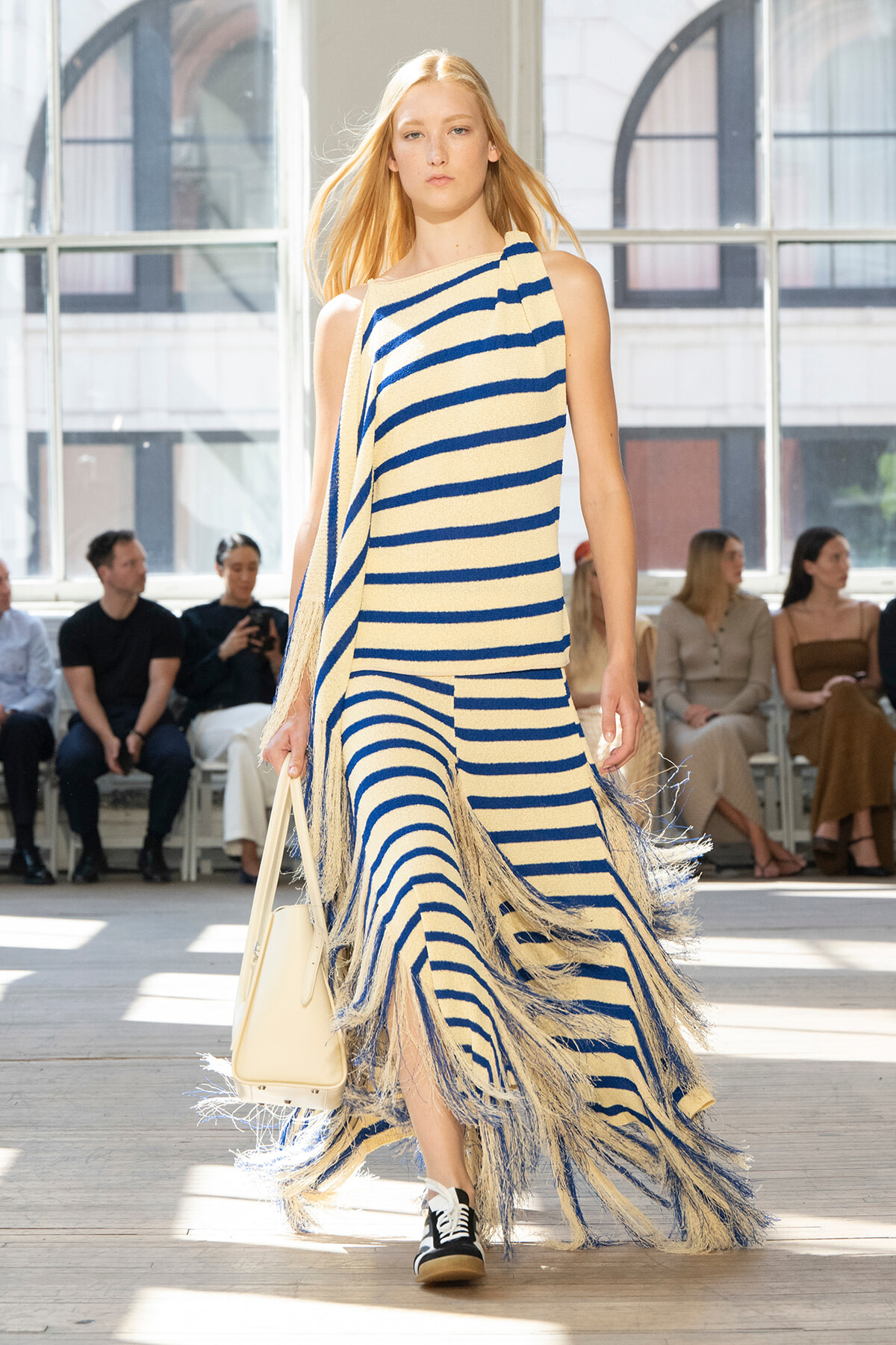 Model walks a cream-and-navy striped fringed dress down a runway in a bright studio, carrying a cream handbag.