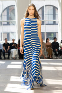 Model walks the runway in a blue and white striped fringe dress, with an audience seated in a bright studio backdrop.