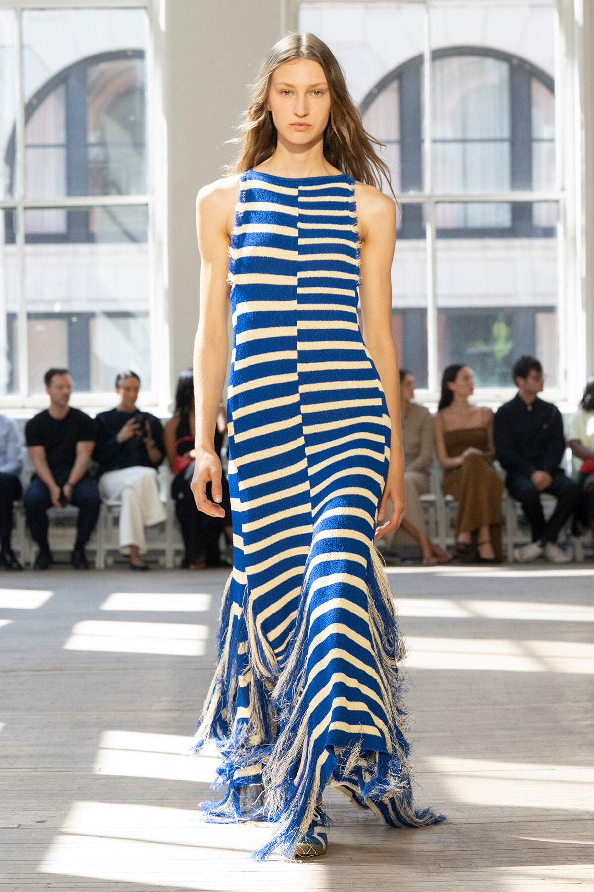 Model walks the runway in a blue and white striped fringe dress, with an audience seated in a bright studio backdrop.