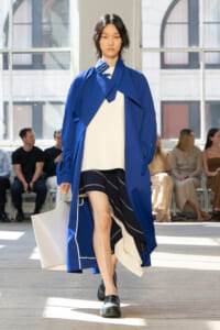 Model walking a fashion runway in a cobalt blue oversized coat over a white top and black striped skirt, carrying a white tote bag.
