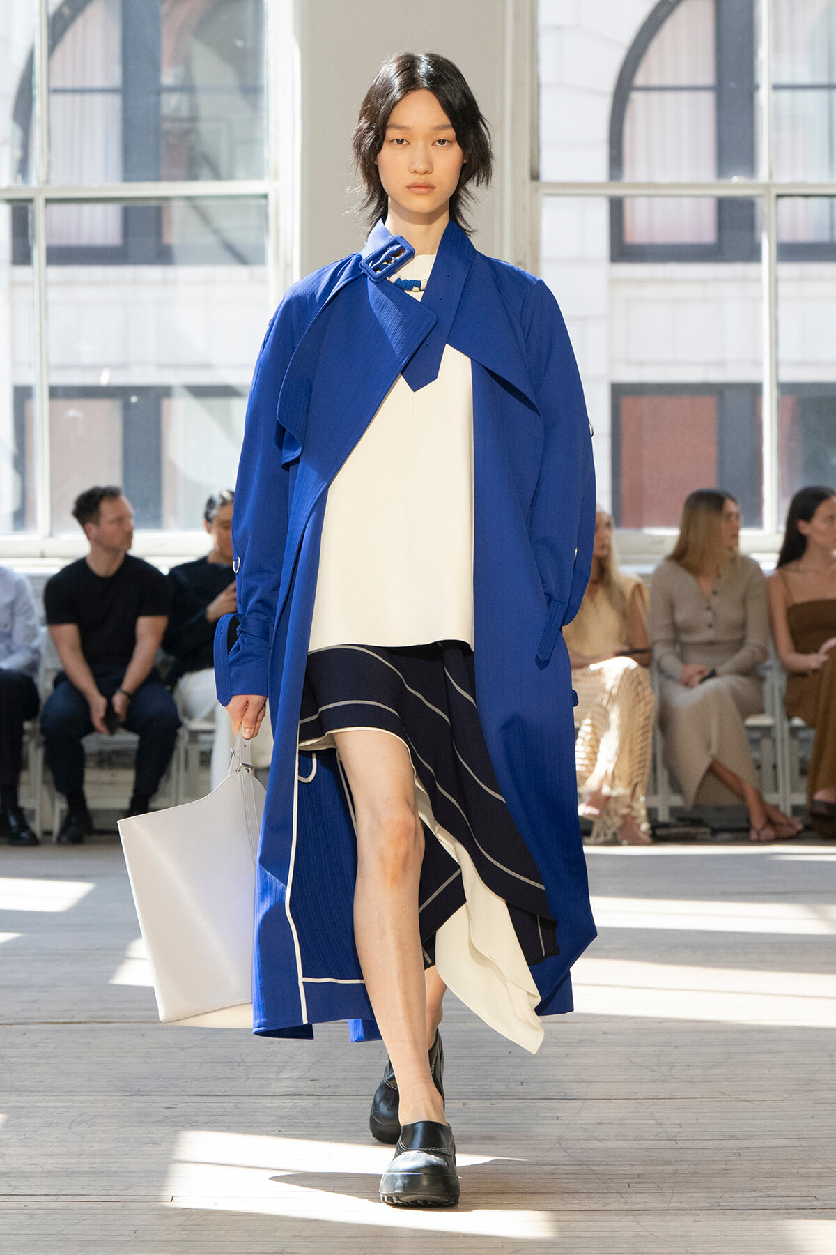 Model walking a fashion runway in a cobalt blue oversized coat over a white top and black striped skirt, carrying a white tote bag.