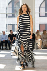 Model walks a runway in a black-and-white striped sleeveless dress with a fringed, layered hem and matching scarf.