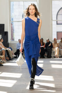 Model walks down a runway in a vivid blue one-shoulder dress with a front knot, holding a white clutch.