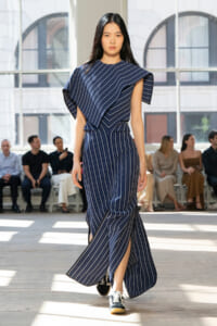 Model walking a runway in a navy blue pinstripe dress with a dramatic draped top.
