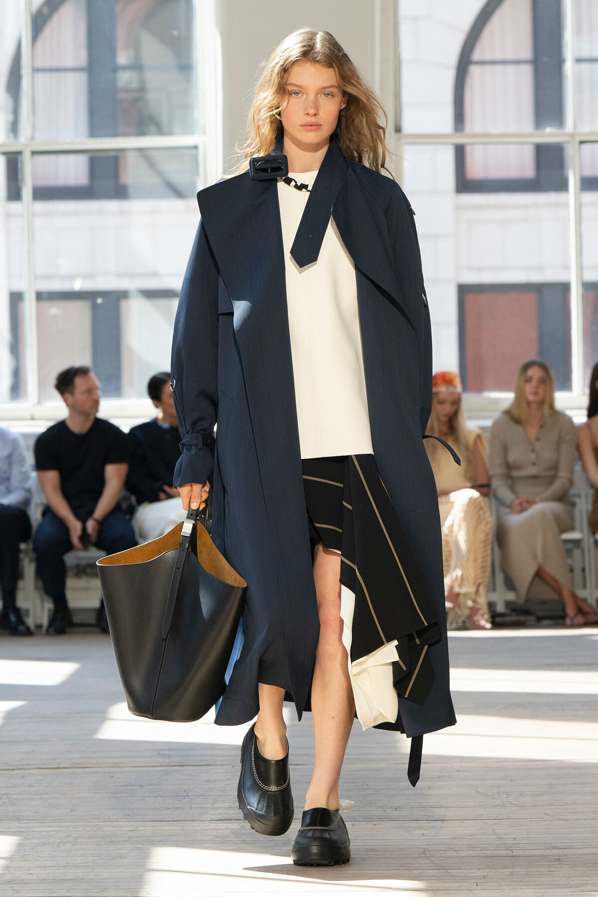 Model walks the runway in a navy oversized trench over a white top and black asymmetrical skirt, carrying a large black tote.