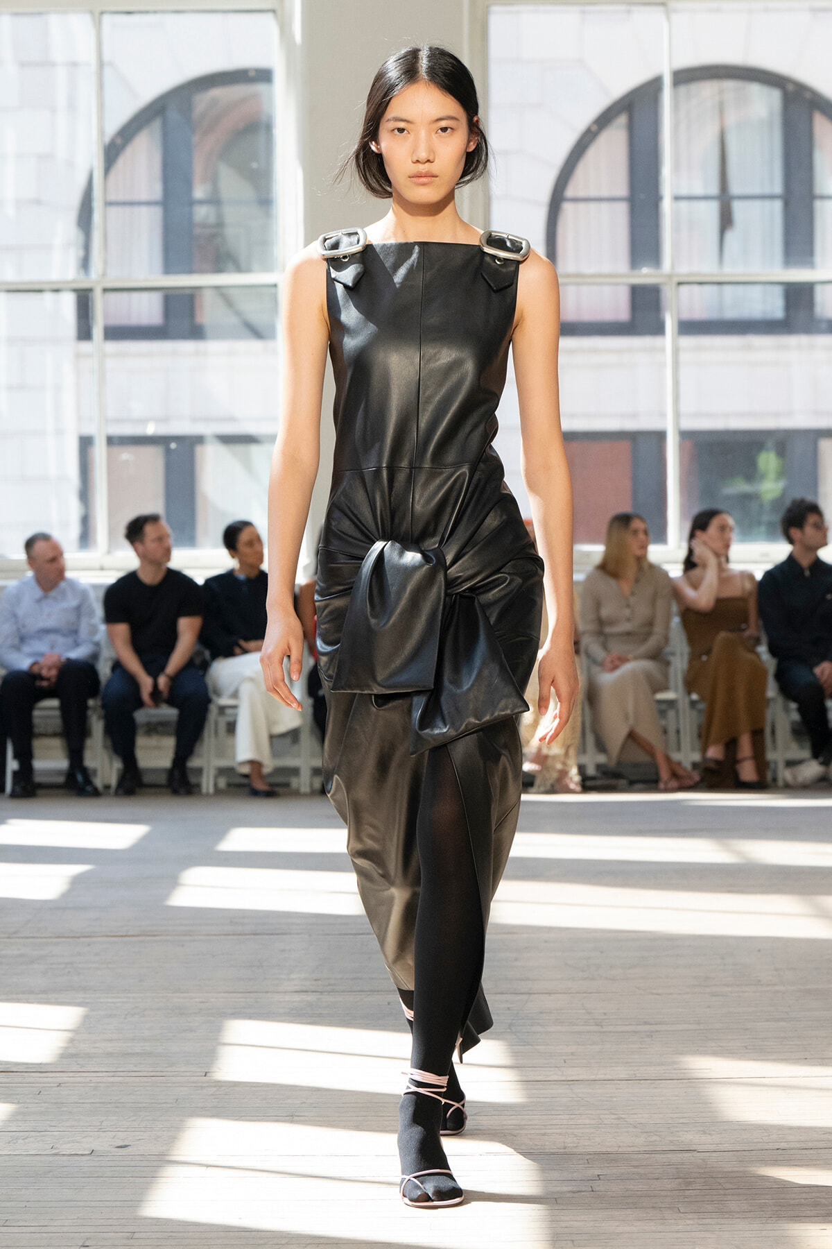Model walks a runway in a sleeveless black leather dress with a large front knot and draped detail; audience sits in the background.