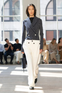 Model walks a fashion runway in a navy pinstriped double-breasted blazer and white wide-leg pants, carrying a small dark blue cylindrical bag.