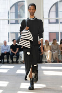 Model walking a fashion runway in a black outfit, carrying a black-and-white striped knit blanket and bun-smash coat draped over her arm, in a bright studio setting.
