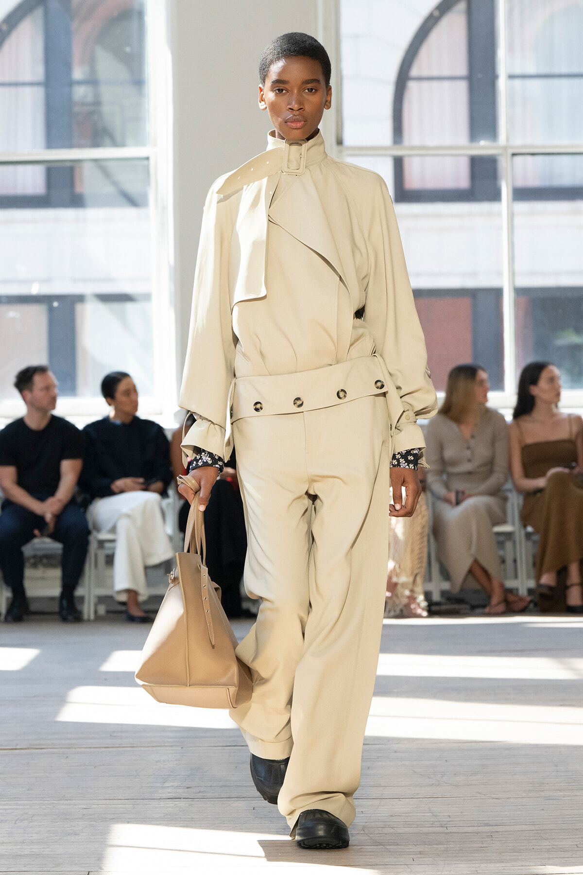Model walks a beige oversized trench with belt and matching wide-leg pants, carrying a tan handbag during a fashion show.