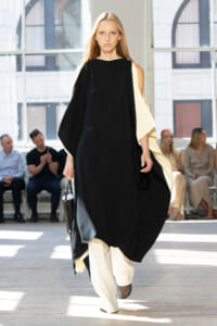 Model walking a runway in a black draped dress with cream cape sleeves and a small black handbag, blond hair flowing.