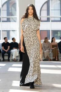 Female model walking a fashion show wearing a long beige floral-print dress with a black panel on the side, in a bright studio with seated spectators behind her.