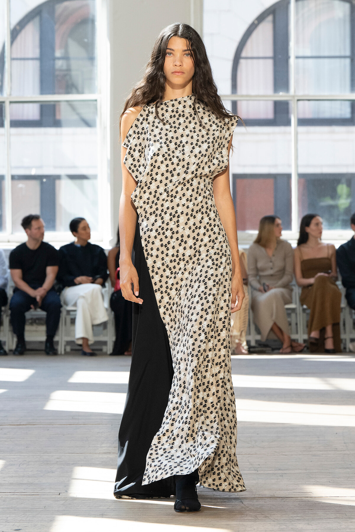 Female model walking a fashion show wearing a long beige floral-print dress with a black panel on the side, in a bright studio with seated spectators behind her.