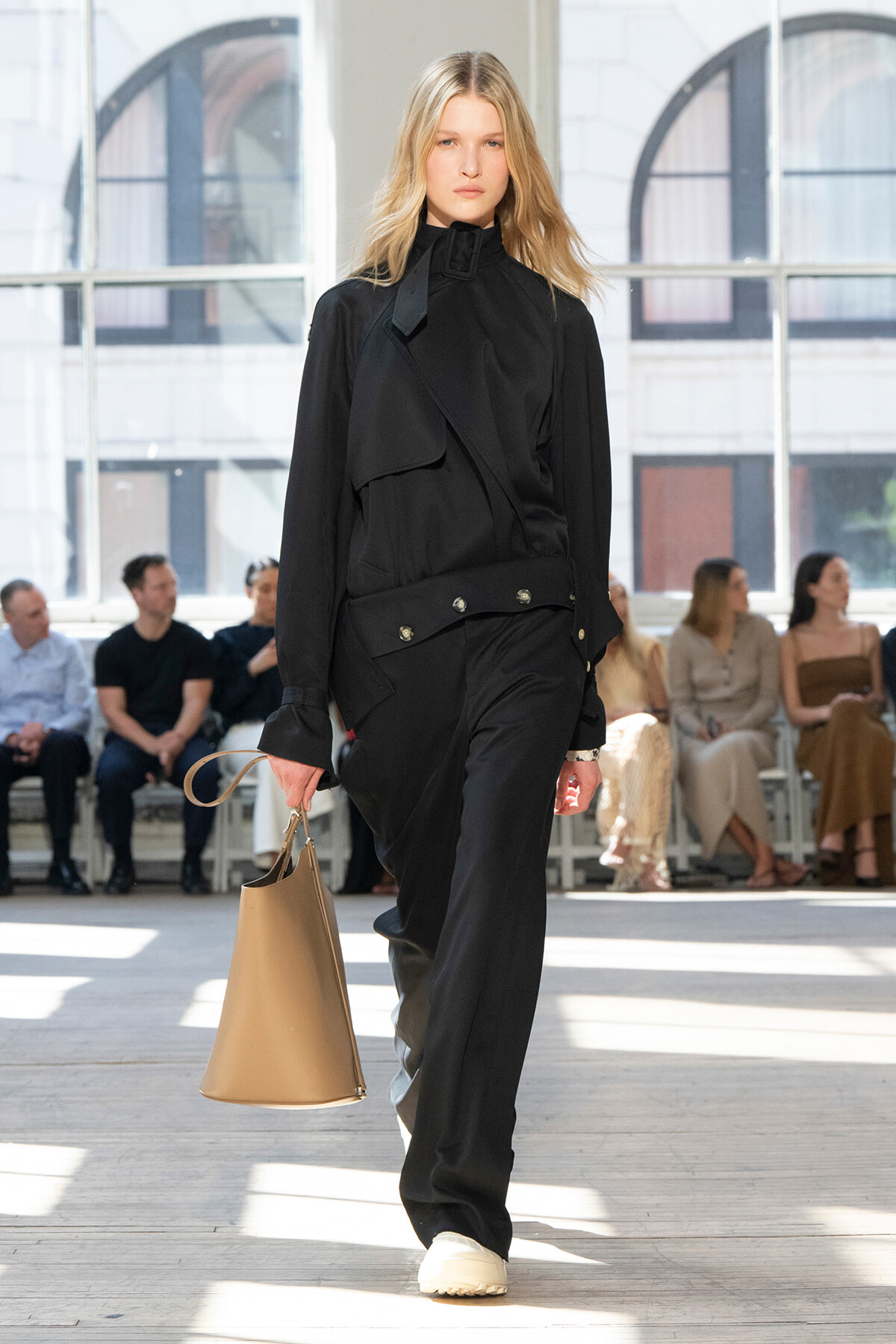 Model walks a runway in a black layered utility outfit with a beige tote bag tucked in hand and sunlit studio backdrop behind audience members.