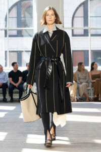 Model wearing a black striped coat with a wide belt walking a fashion runway, holding a beige bag.