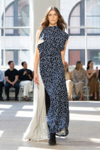 Model walking the runway in a navy floral dress with a white cape-like back detail, audience watching in a bright venue