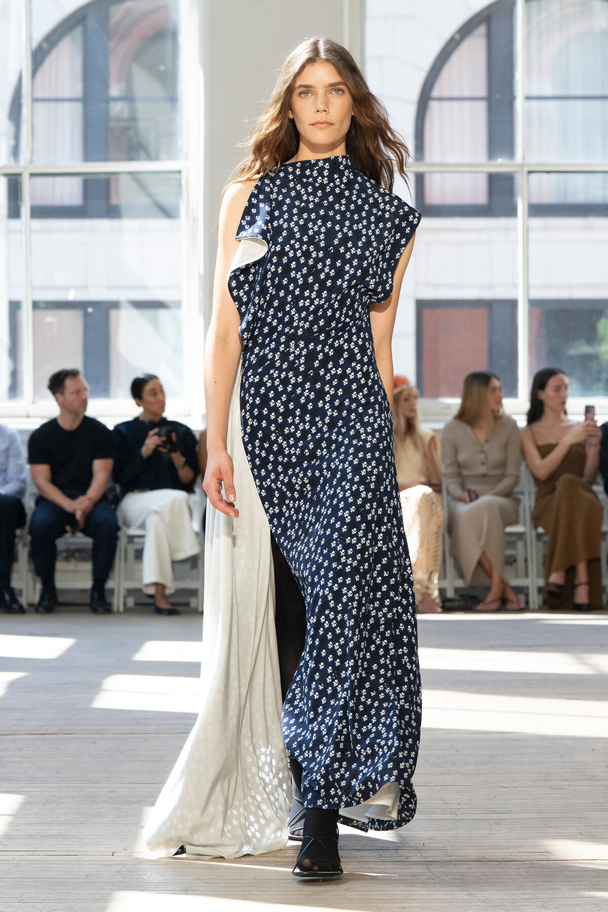 Model walking the runway in a navy floral dress with a white cape-like back detail, audience watching in a bright venue