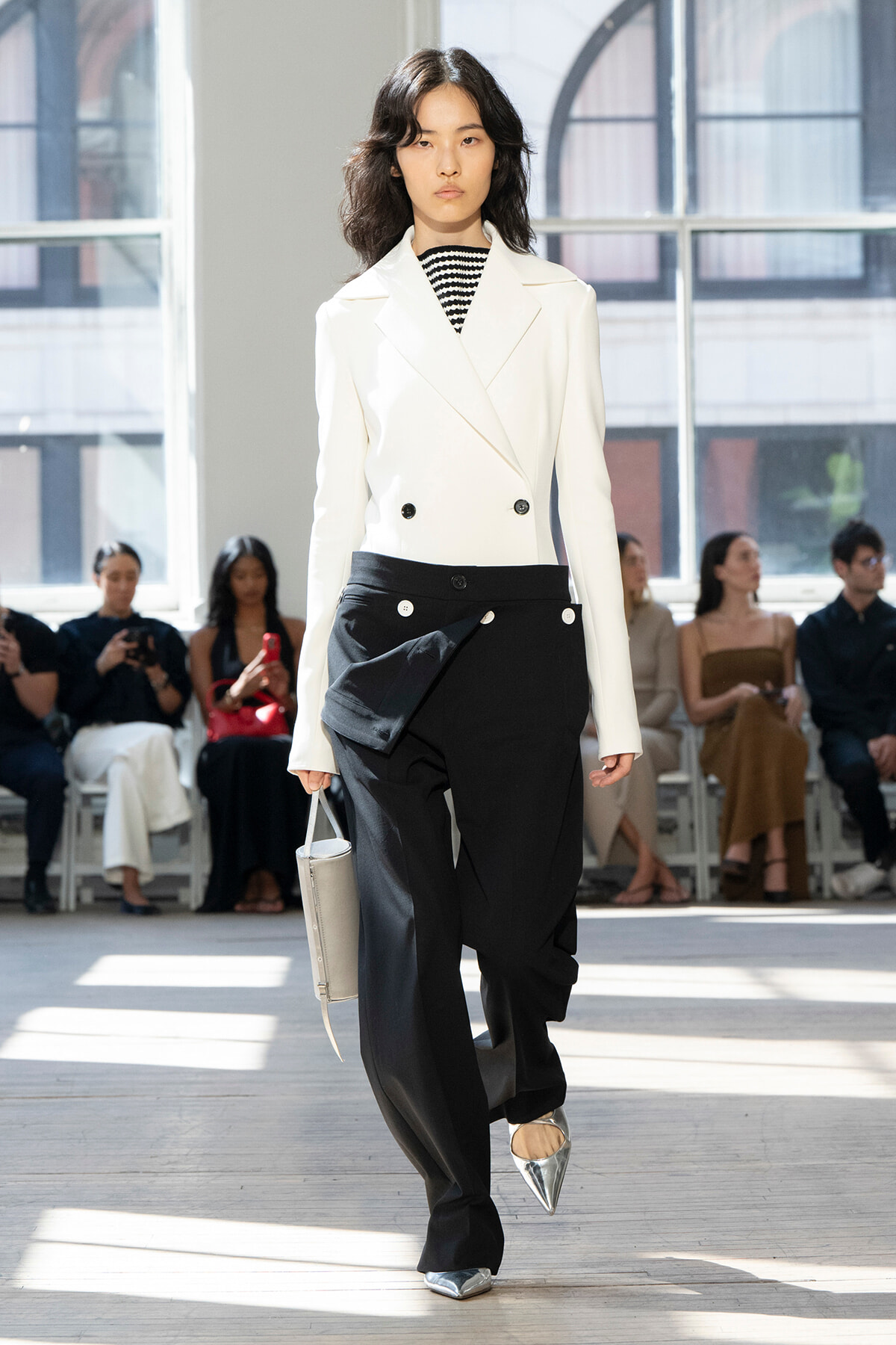 Runway model in a white double-breasted blazer and black wide-leg pants with draped panels, wearing silver pointed flats and carrying a light gray handbag.