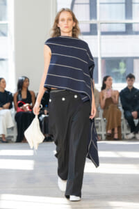 Model on a runway wearing a navy striped poncho-style top and black wide-leg pants, holding a white bag.