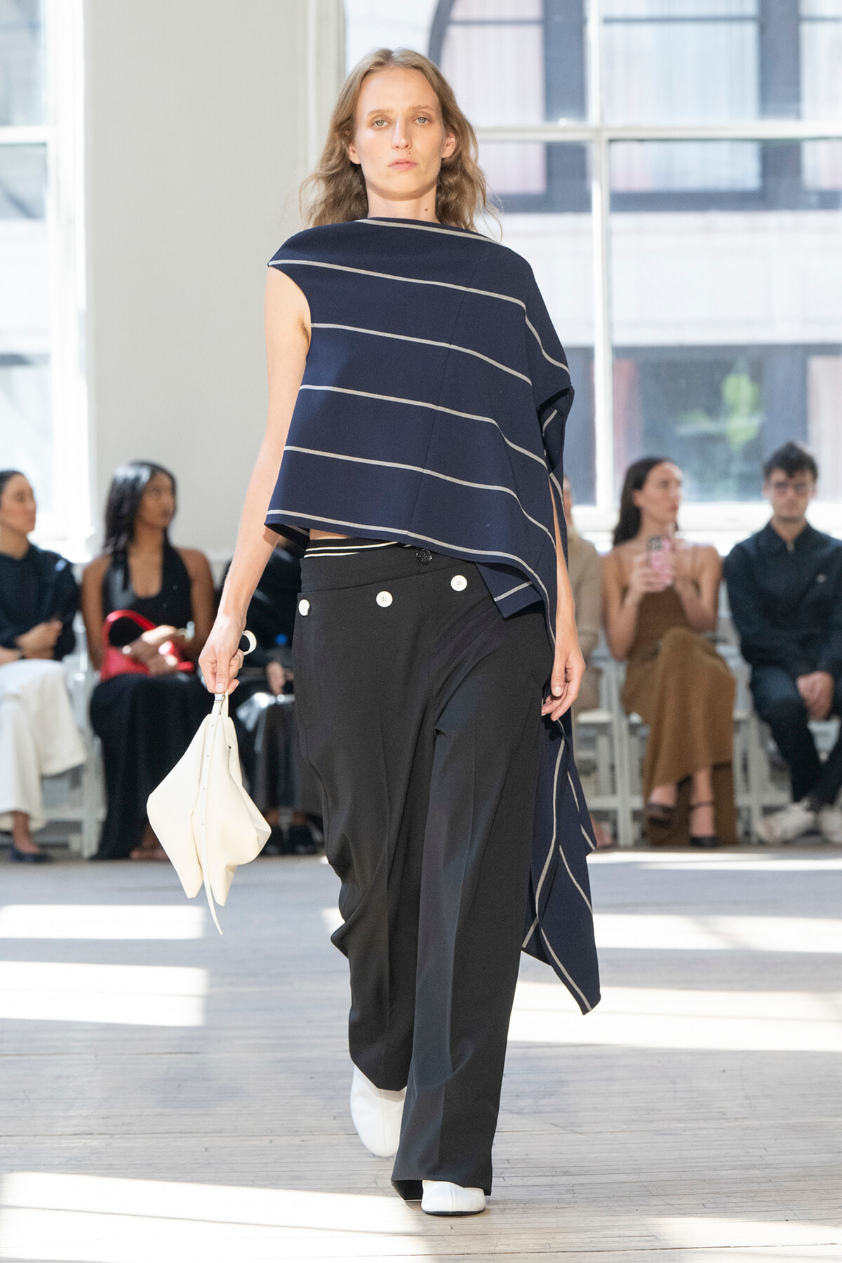 Model on a runway wearing a navy striped poncho-style top and black wide-leg pants, holding a white bag.