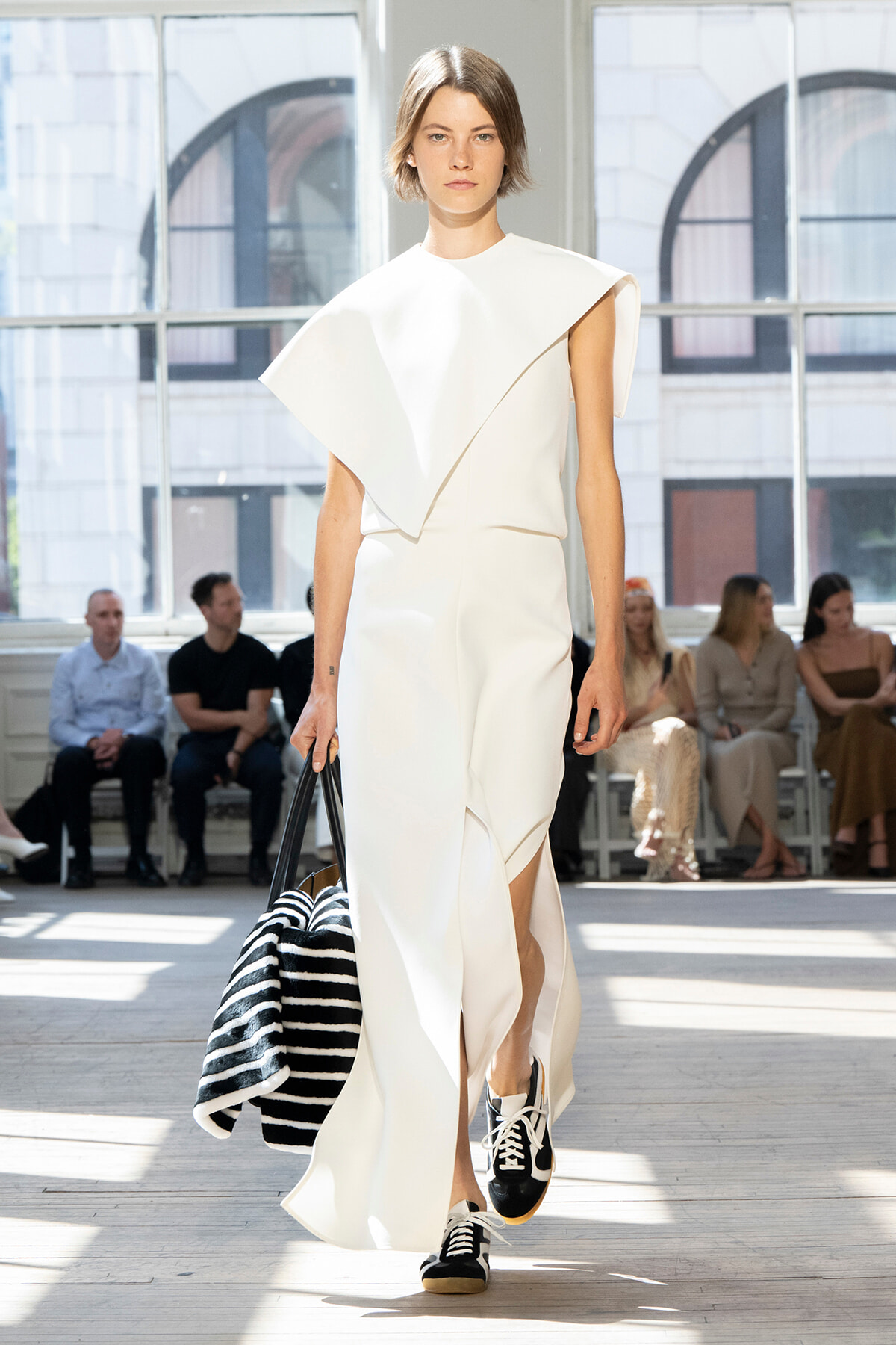 Runway model in a white sculptural dress with a cape-like detail, carrying a black-and-white striped bag.