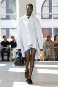 Model walks the runway in an oversized light blue striped shirt dress, carrying a black handbag, with feathered sandals and a choker; audience seated in the background.
