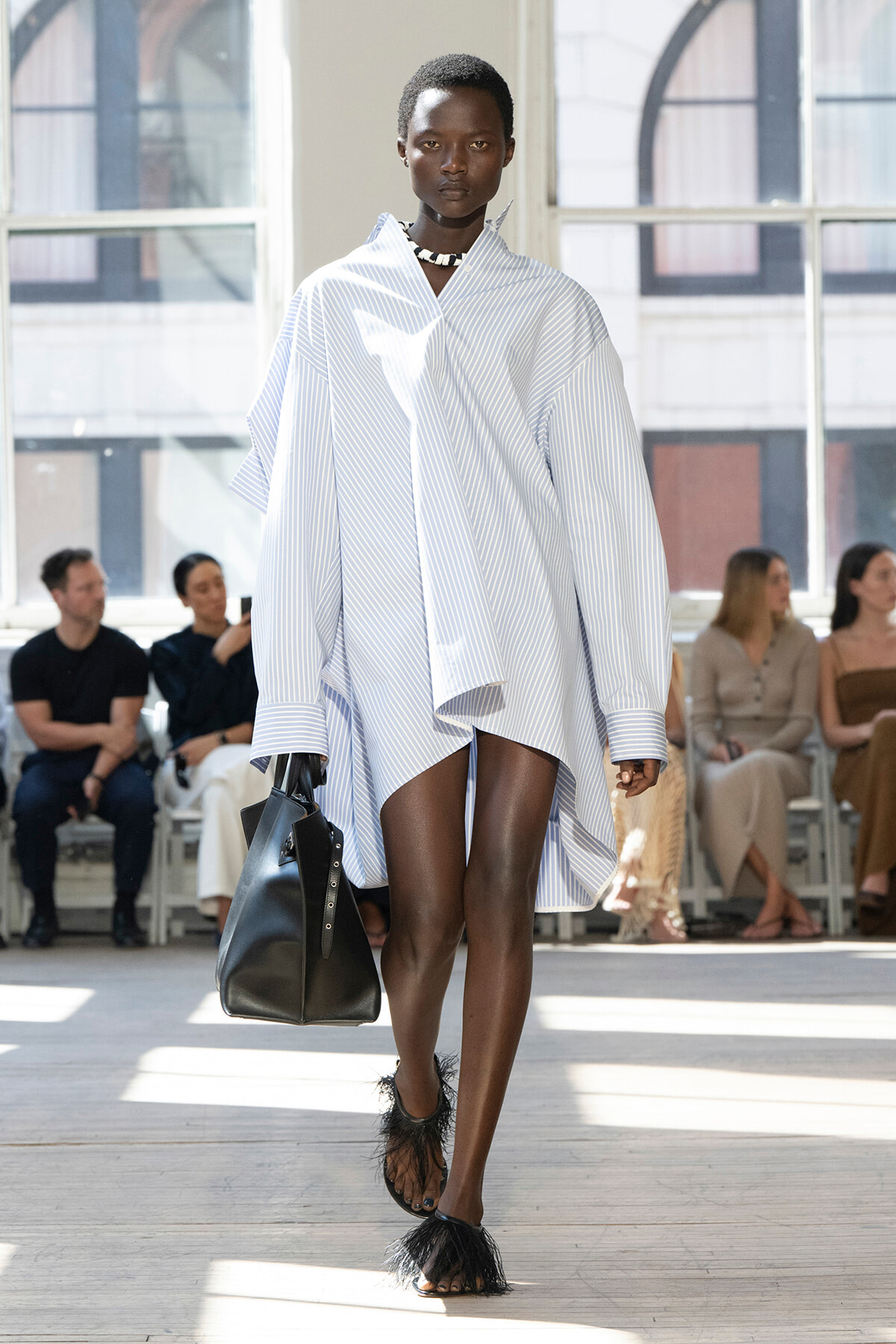 Model walks the runway in an oversized light blue striped shirt dress, carrying a black handbag, with feathered sandals and a choker; audience seated in the background.