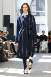 Model walking a runway in a navy pinstriped coat with a black belt, paired with a striped shirt and pointed heels.