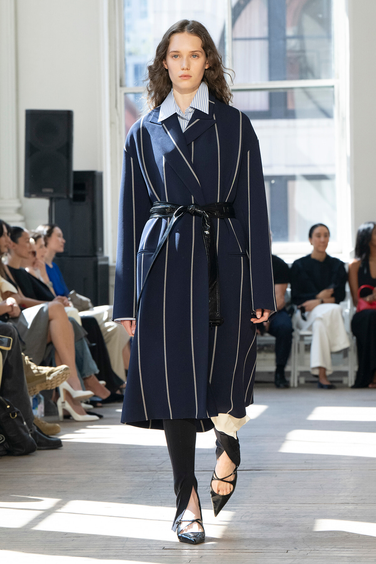 Model walking a runway in a navy pinstriped coat with a black belt, paired with a striped shirt and pointed heels.