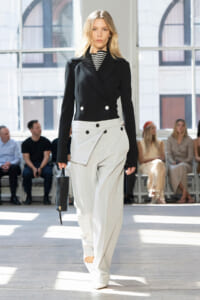 Female model walks a fashion show in a black double-breasted blazer and light gray wide-leg pants with a large front pocket, holding a small black bag, audience in background.