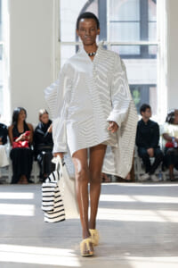 Model walking the runway in an oversized white shirt dress with blue stripes and cape-like sleeves, carrying a white striped tote with a striped scarf, and feathered-trim sandals.