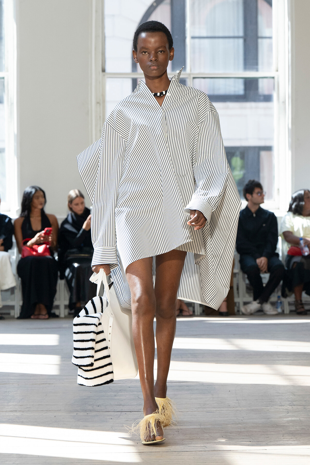 Model walking the runway in an oversized white shirt dress with blue stripes and cape-like sleeves, carrying a white striped tote with a striped scarf, and feathered-trim sandals.