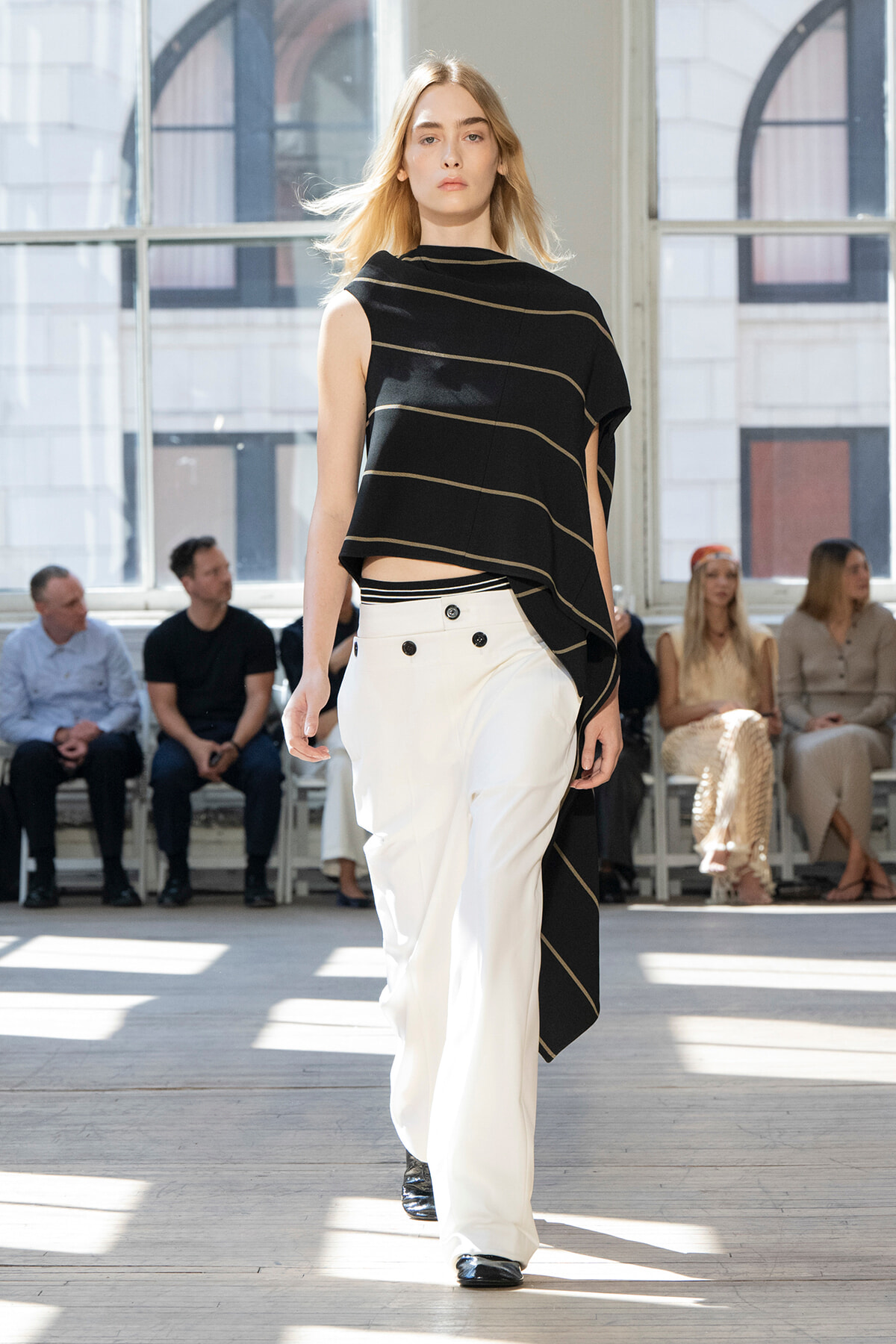 Model on a fashion runway wearing a black striped asymmetrical top and white wide-leg pants with black shoes, audience watching in back.