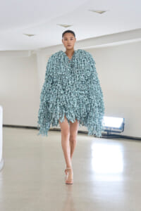 Model walking a runway in a voluminous light-blue cape made of leaf-like fringe pieces.