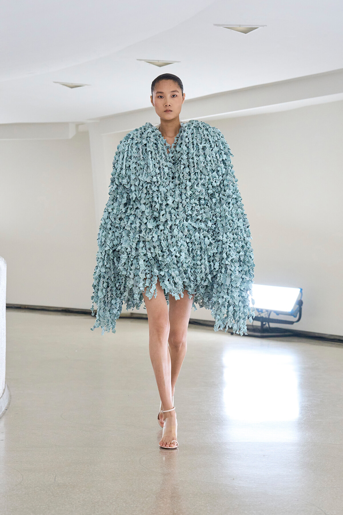Model walking a runway in a voluminous light-blue cape made of leaf-like fringe pieces.