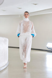 Runway model in a white open-knit mesh dress with a high neck, blue cuffs, and sheer lattice fabric in a bright studio.
