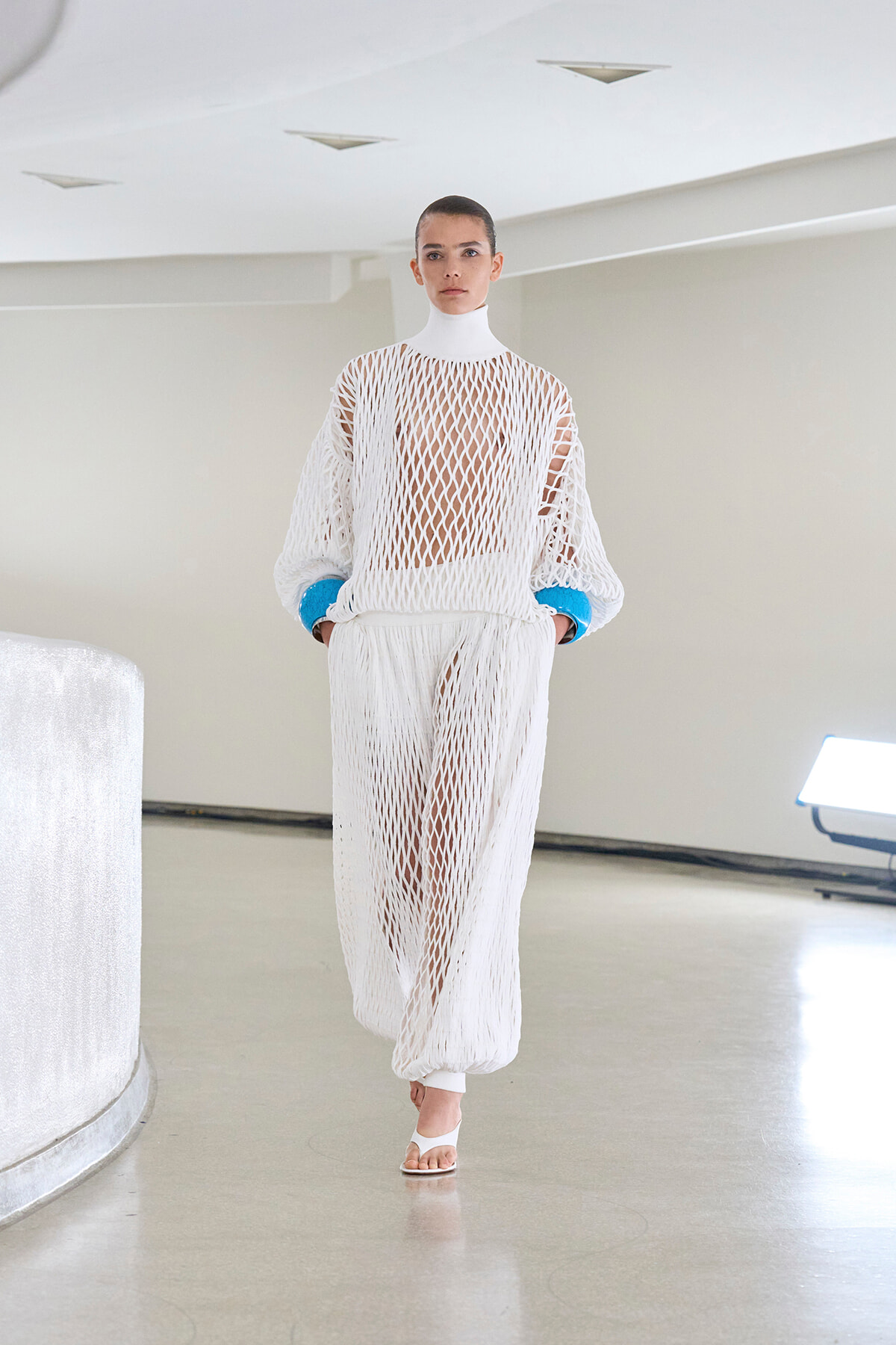 Runway model in a white open-knit mesh dress with a high neck, blue cuffs, and sheer lattice fabric in a bright studio.