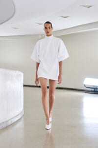 Model wearing a white oversized mini dress with a high neckline and wide sleeves, paired with white heeled mules, walking in a bright studio.