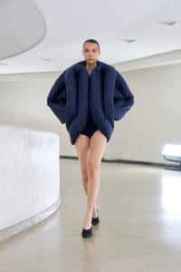 Model walking in a stark white space wearing a sculptural navy puffer garment that envelops the torso, with black heels visible.