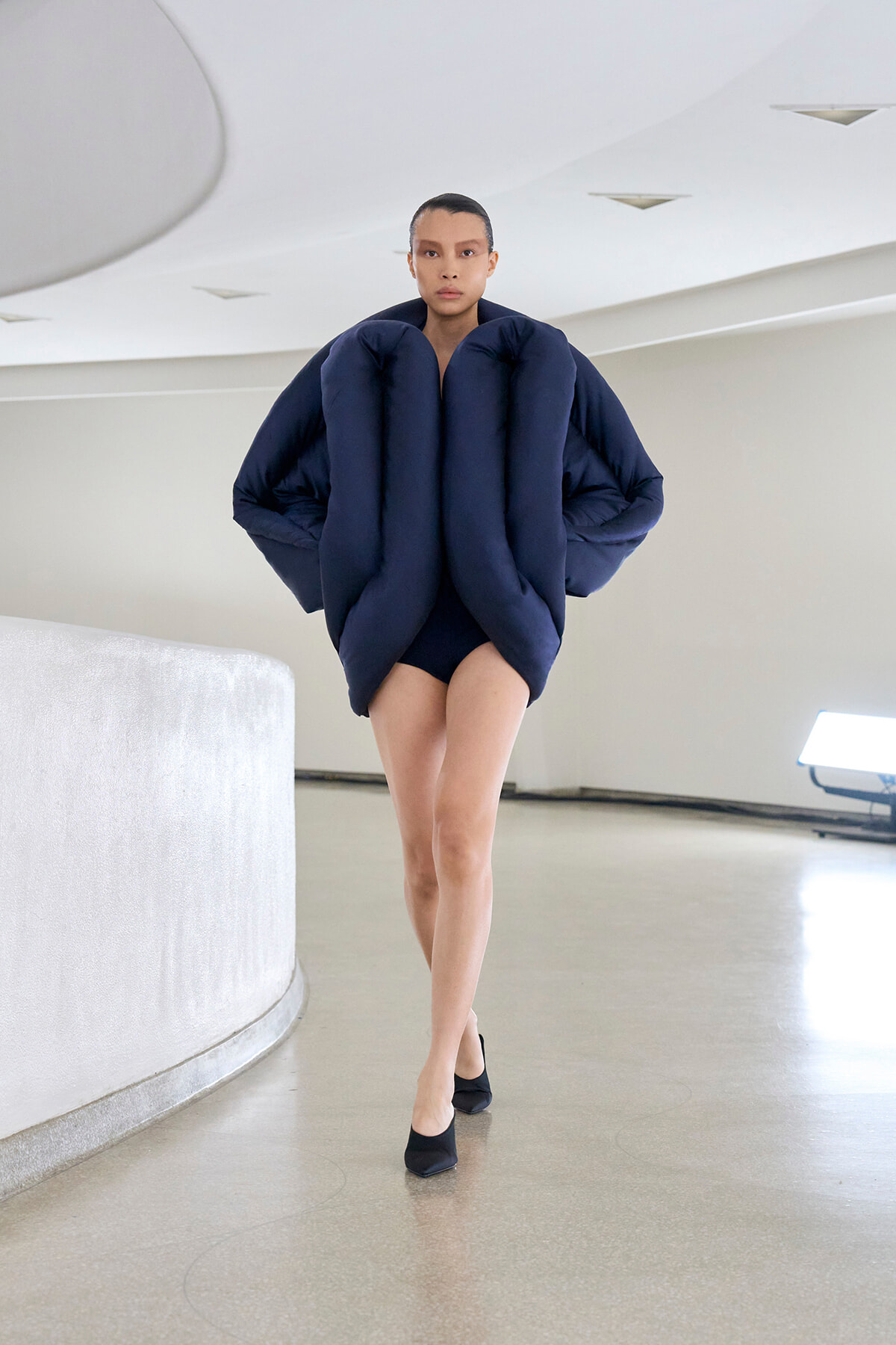 Model walking in a stark white space wearing a sculptural navy puffer garment that envelops the torso, with black heels visible.