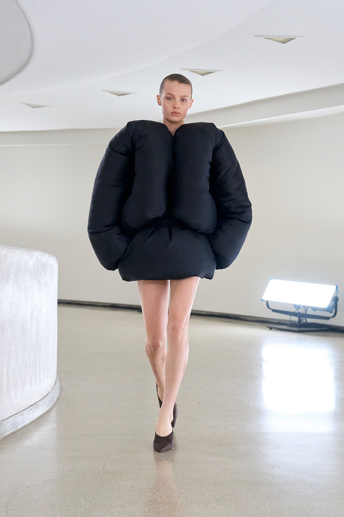 Model walks a minimalist runway wearing an oversized black puffer dress with dramatically padded shoulders and burgundy heels.