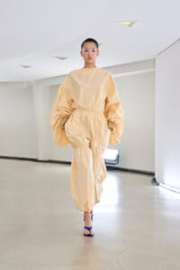 Female model walks down a minimalist runway wearing a peach oversized jumpsuit with gathered waist and balloon sleeves, blue sandals visible at feet.