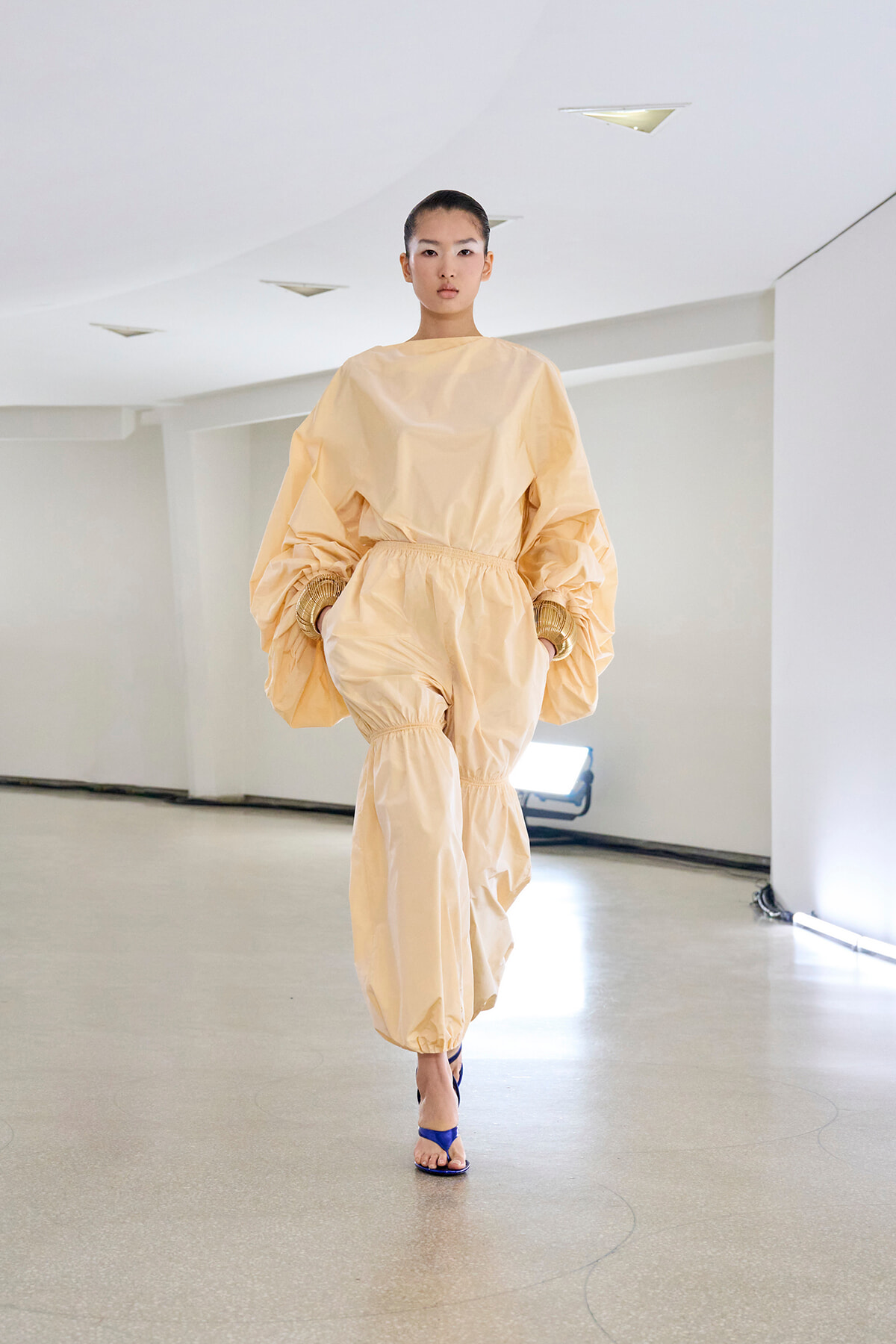 Female model walks down a minimalist runway wearing a peach oversized jumpsuit with gathered waist and balloon sleeves, blue sandals visible at feet.