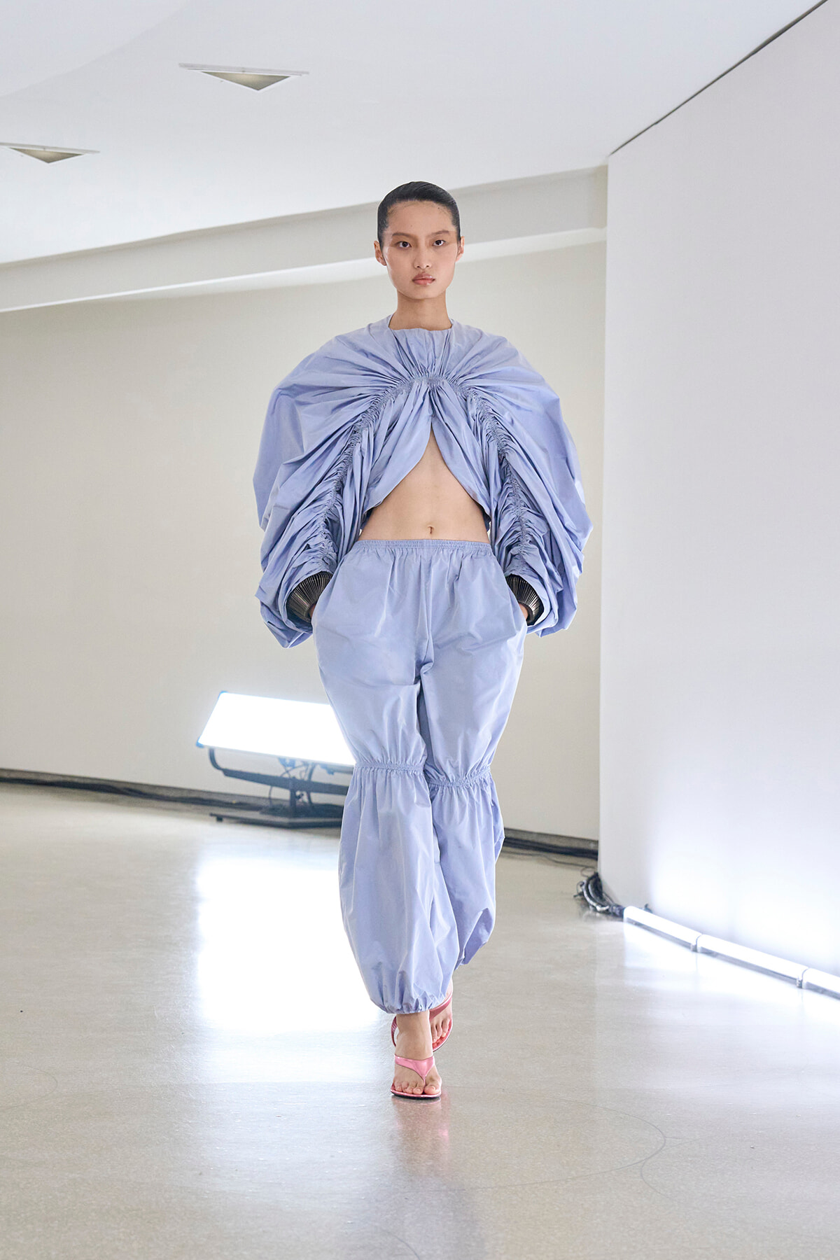 Model on a runway wearing a lavender-blue sculptural top with ruching at the center and matching loose pants, midriff exposed, pink sandals visible.