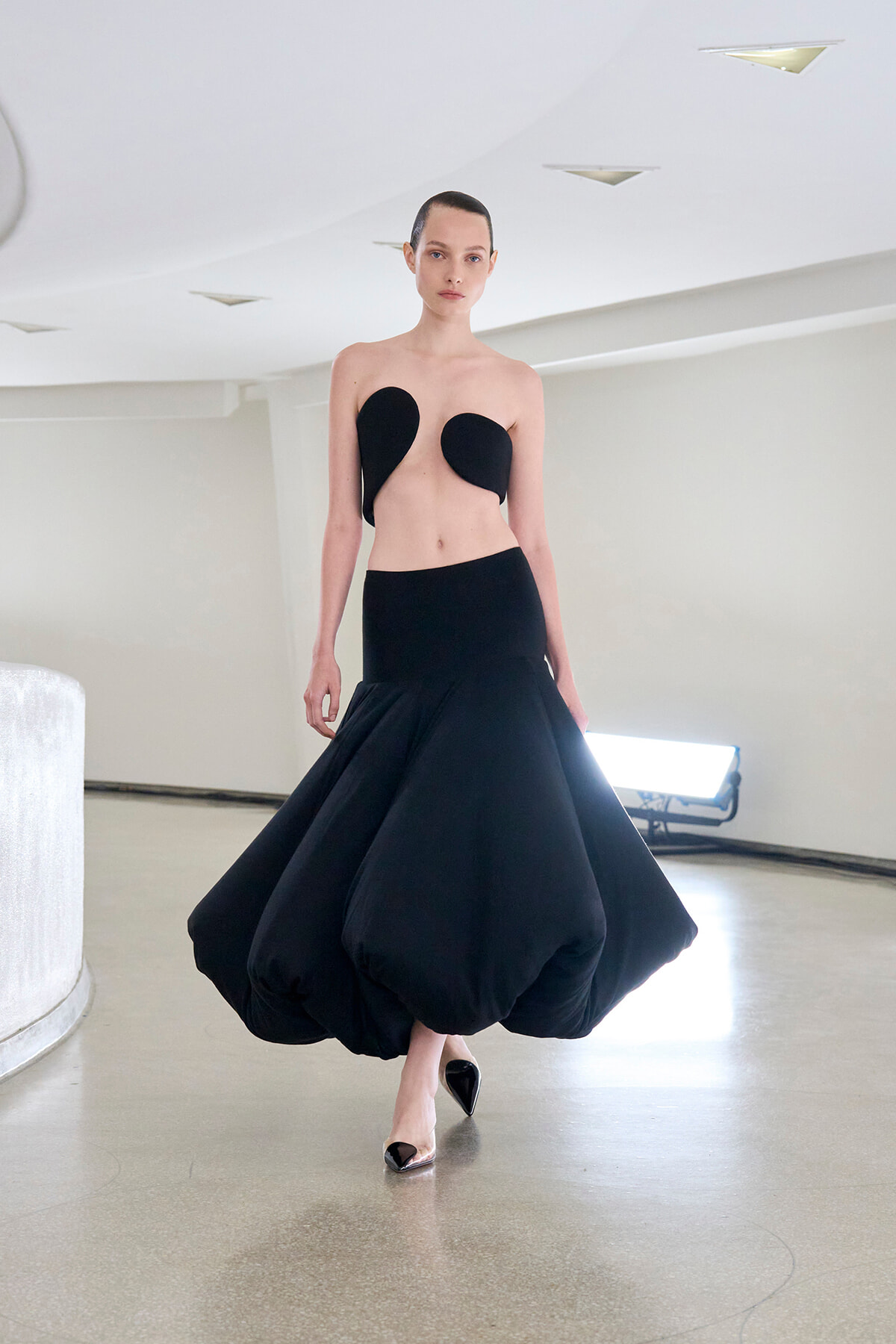 Runway model in a minimalist black sculptural outfit: strapless top with curved cups and a voluminous bubble skirt.