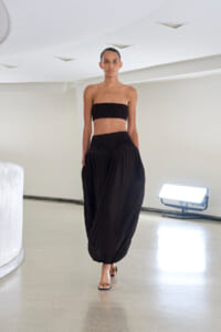 Model walking in a white studio wearing a black strapless bandeau top and matching wide, gathered pants with sandals shift-heels.