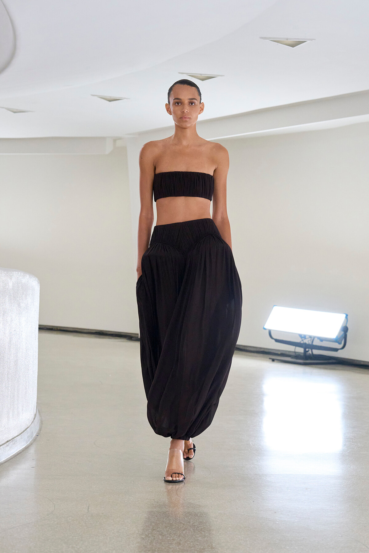 Model walking in a white studio wearing a black strapless bandeau top and matching wide, gathered pants with sandals shift-heels.