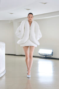 Model wearing an oversized white puffer garment with large looped folds, walking in a bright, minimalist space.