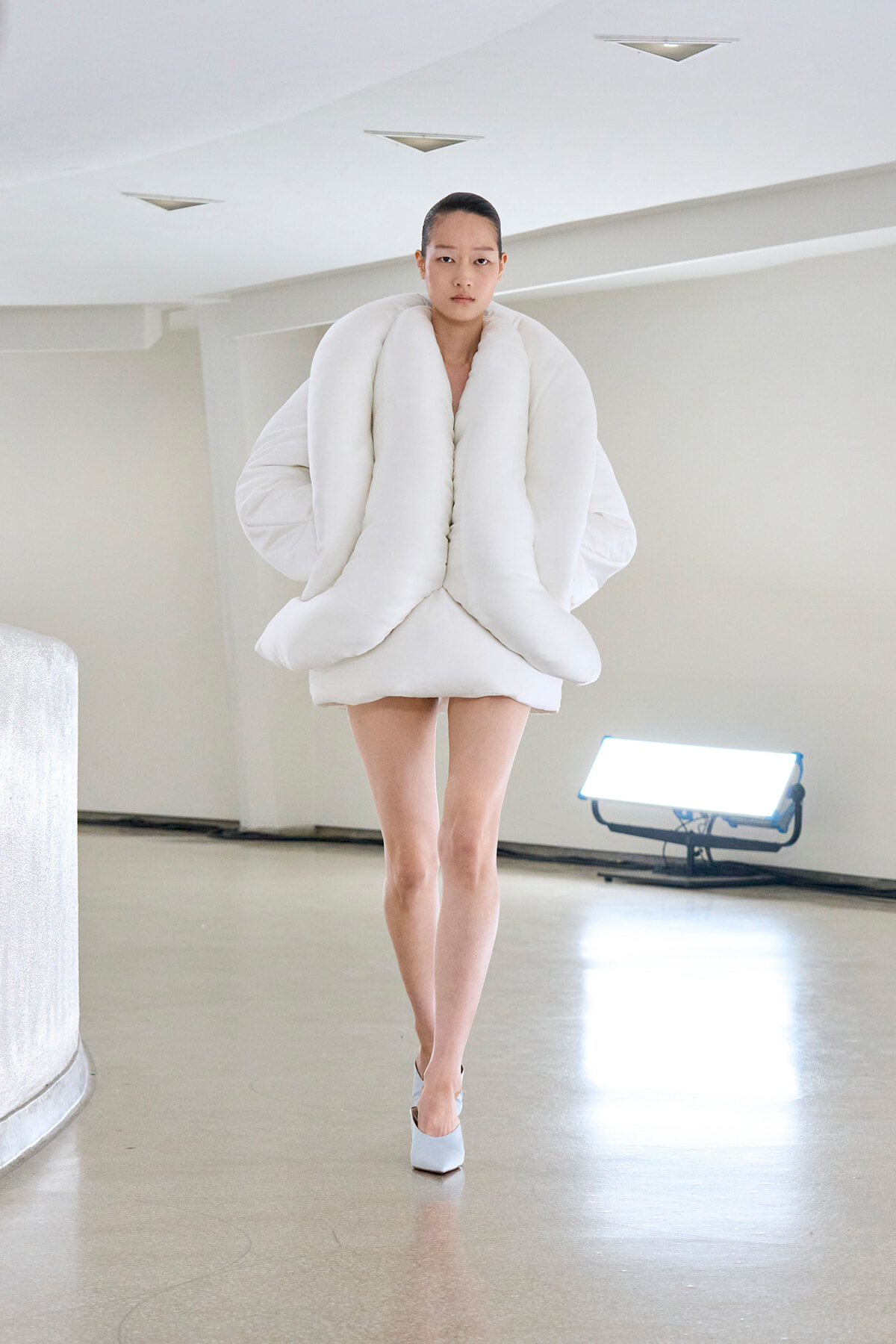 Model wearing an oversized white puffer garment with large looped folds, walking in a bright, minimalist space.
