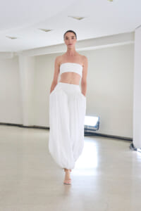 Model walking in a white bandeau top and wide, pleated white pants in a bright studio setting.