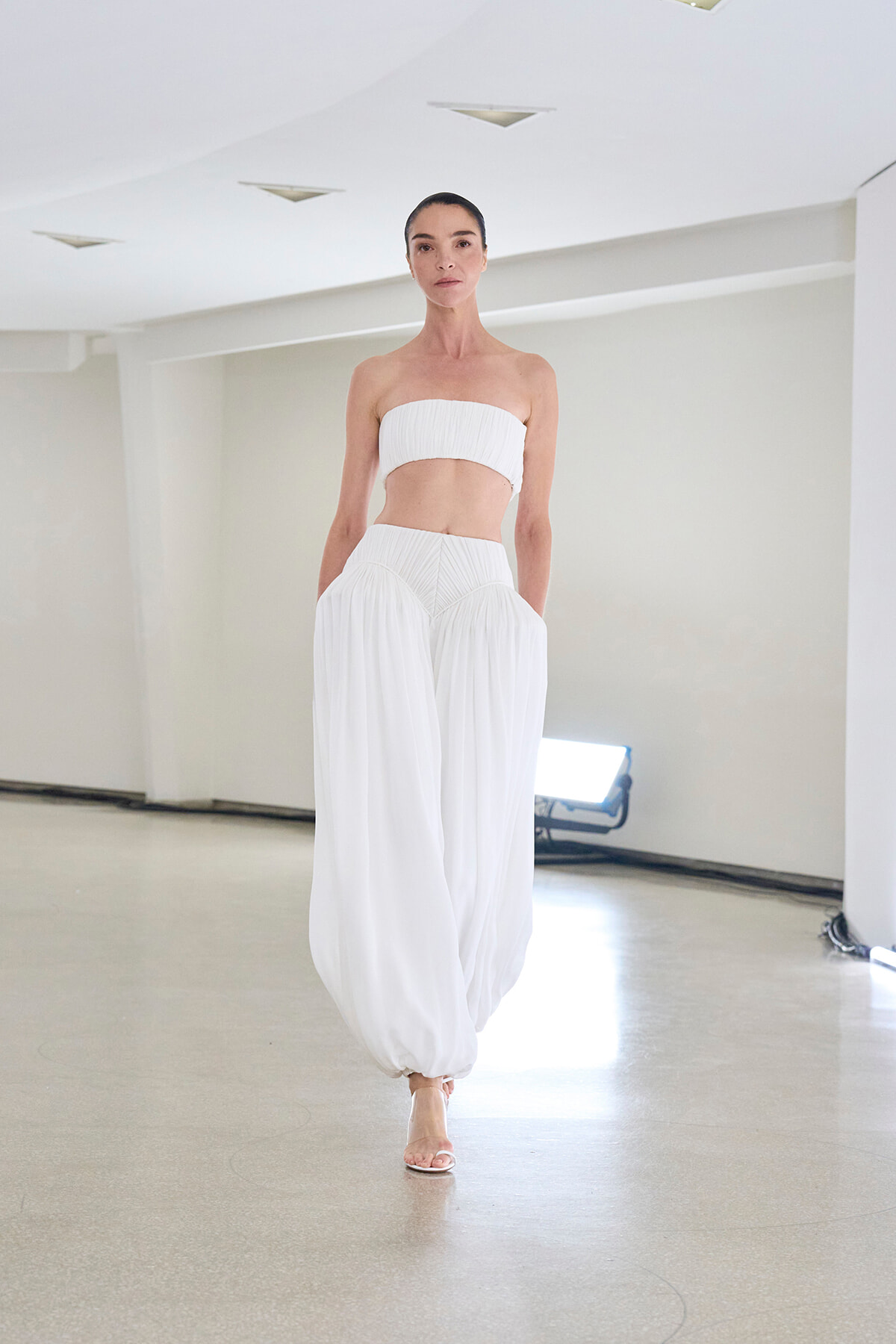 Model walking in a white bandeau top and wide, pleated white pants in a bright studio setting.